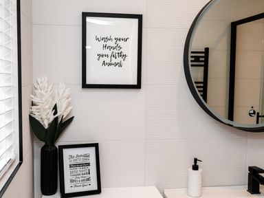 A bathroom wall with two framed signs a vase with white flowers a round mirror and a soap dispenser at Polished Pebble in Beadnell