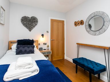 A bedroom with a single bed with white and blue bedding and towels on top a wooden door a bedside table with a lamp a round wall mirror a blue bench and wall hooks at Polished Pebble in Beadnell