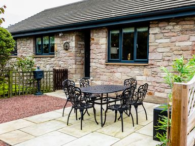 An outdoor patio area with a metal table and six chairs next to a stone wall house at Polished Pebble in Beadnell