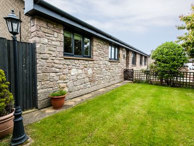 A stone wall house with black framed windows and a green lawn with potted plants and a lamp post at Polished Pebble in Beadnell