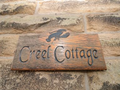 A wooden sign with a lobster carving and Creel Cottage text mounted on a stone wall at Creel Cottage Blackwood Street in Amble