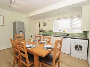 A kitchen with wooden dining table set for six with plates and glasses near a refrigerator sink and washing machine at Creel Cottage Blackwood Street in Amble