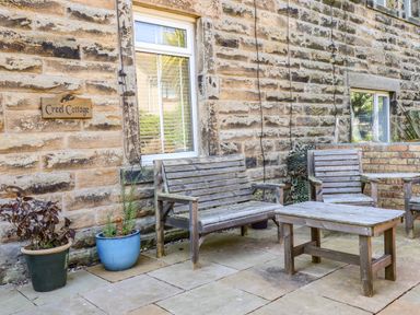 An outdoor seating area with wooden benches and table next to a stone wall with a window and potted plants at Creel Cottage Blackwood Street in Amble