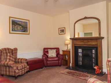 A sitting room with armchairs a fireplace a wooden side table with a lamp and framed pictures on the walls at Creel Cottage Blackwood Street in Amble