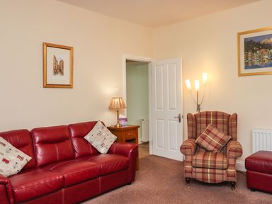 A living room with a red leather sofa, a plaid armchair with a cushion, a side table with a lamp, an open door, and framed pictures at Creel Cottage Blackwood Street in Amble