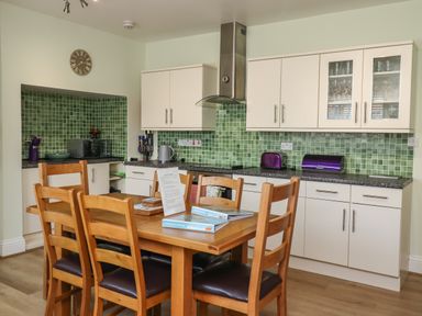 A kitchen with wooden dining table and chairs white cabinets green tiled backsplash and various appliances at Creel Cottage Blackwood Street in Amble