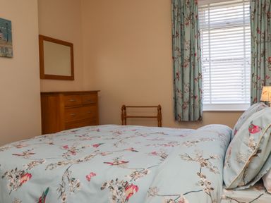 A bedroom with a bed with floral bedding a wooden dresser with mirror a window with blinds and floral curtains at Creel Cottage Blackwood Street in Amble