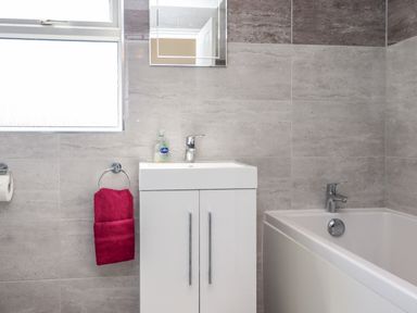 A bathroom with a white sink cabinet a red towel on a ring a mirror a window and a bathtub at Creel Cottage Blackwood Street in Amble