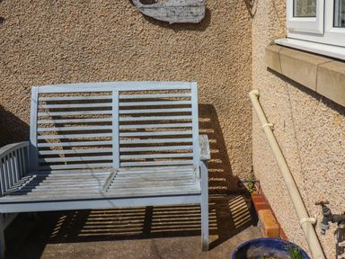 A wooden outdoor bench against a textured wall with a pipe and a window at Creel Cottage Blackwood Street Amble