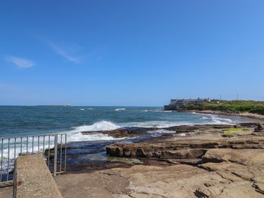 A rocky beach with waves crashing a metal railing a row of houses on a grassy coastline and an island with a lighthouse in the sea at Creel Cottage Blackwood Street in Amble