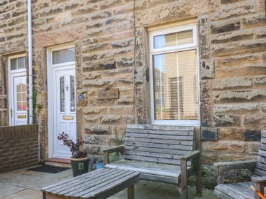 The front entrance of Creel Cottage with a white door wooden outdoor bench table and chair on a stone patio at Creel Cottage Blackwood Street in Amble
