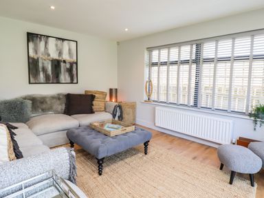 A living room with a grey sectional sofa a tufted ottoman and a large window with blinds at Willow in Beadnell