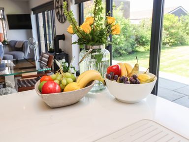 A kitchen countertop with two bowls of fruit and a vase of yellow flowers near a glass dining table and large windows at Willow Beadnell