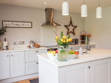 A kitchen with white cabinets an island with a vase of yellow flowers fruit bowls a stainless steel hood and hanging lights at Willow Beadnell