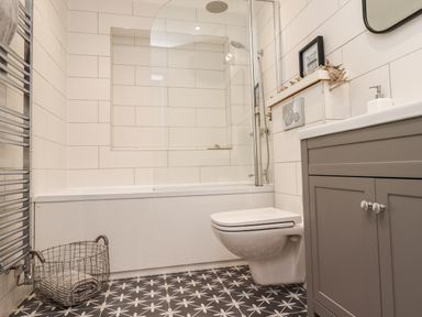 A bathroom with a bathtub shower combination a wall-mounted toilet a grey cabinet and a wire basket on a patterned floor at Willow in Beadnell