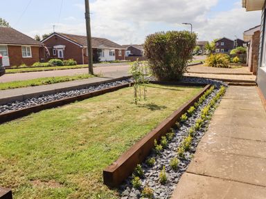 A front garden with grass bordered by stones shrubs and a small tree with houses and a street in the background at Willow in Beadnell