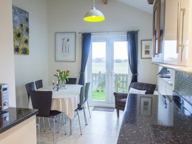 A dining area with a round table and chairs next to glass doors with curtains and a small armchair at Our Story Cottage in Longhoughton