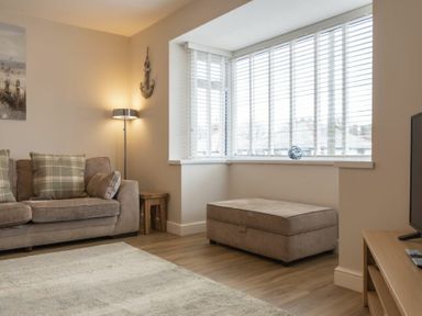 A living room with a sofa a floor lamp an ottoman a television and a bay window with blinds at Southfield Lodge in Seahouses