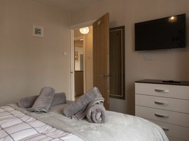 A bedroom with a bed topped with rolled towels a dresser with a television mounted above it and an open door revealing a hallway at Southfield Lodge in Seahouses