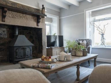 A living room with a wood burning stove a wooden coffee table with cups and pastries and a grey armchair by a window at Creel Cottage Craster