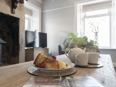 A table with a plate of scones and strawberries and a teapot with cups in a living room with a television and plants at Creel Cottage Craster
