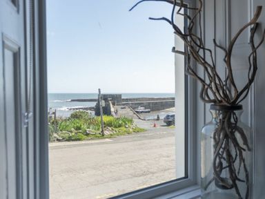 A view of a harbor and ocean through a window with a glass jar holding twigs on the windowsill at Creel Cottage Craster