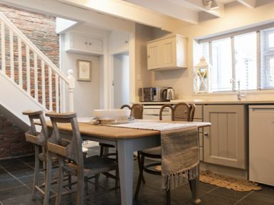 A kitchen with a wooden dining table and chairs near a brick wall and white staircase at Creel Cottage Craster