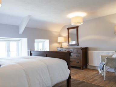 A bedroom with a wooden bed, wooden dresser with mirror, table lamp, white chair with blanket on hardwood floor at Creel Cottage Craster