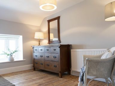 A room with a wooden dresser with a mirror and lamp next to a window and a wicker chair with pillows at Creel Cottage Craster