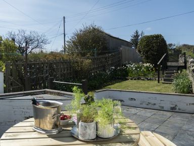A garden with a round wooden table holding plants a metal ice bucket and two glasses by a paved patio area at Creel Cottage Craster