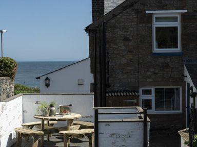 An outdoor seating area with a round wooden table and benches near a stone house by the sea at Creel Cottage Craster