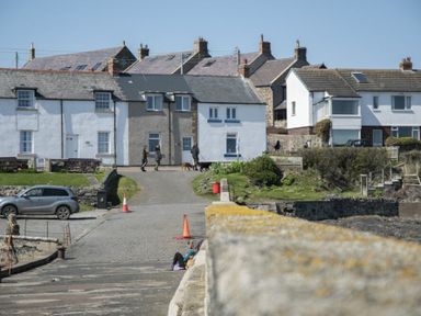 A street with houses and people walking dogs near a parking area at Creel Cottage Craster