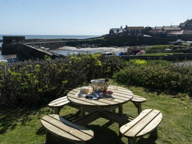 A round wooden picnic table with benches on grass near a harbor and houses at Creel Cottage Craster