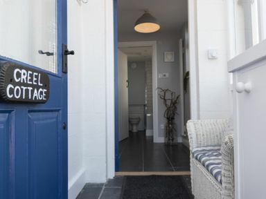 An open blue door with a Creel Cottage sign leading to a hallway with a wicker chair and a pendant light at Creel Cottage Craster