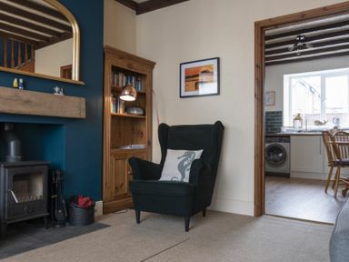 A living room with a black armchair, wooden bookshelf, fireplace, and a view into a kitchen with a washing machine at Kittiwake Cottage Seahouses