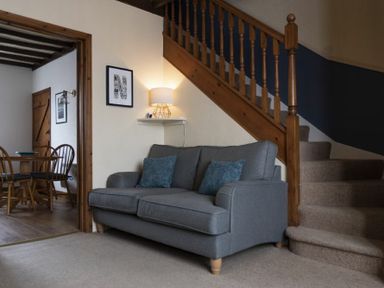 A living room with a gray sofa and cushions next to wooden stairs and a dining room with wooden chairs in the background at Kittiwake Cottage Seahouses