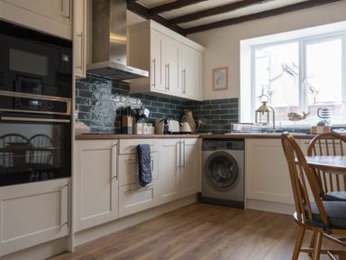 A kitchen with white cabinets a washing machine wooden dining chairs and a window at Kittiwake Cottage Seahouses
