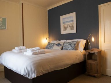 A bedroom with a bed with white linens and towels on it a wooden nightstand with a lamp and a framed picture on a blue wall at Kittiwake Cottage Seahouses