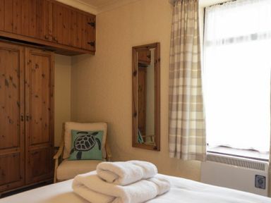 A bedroom with a bed with folded white towels a wooden wardrobe a chair with a turtle cushion a wall mirror and a window with checkered curtains at Kittiwake Cottage Seahouses