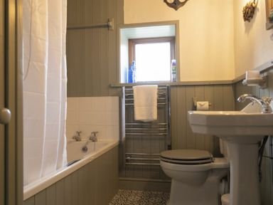 A bathroom with a bathtub with shower curtain a toilet a pedestal sink and a towel rack under a window at Kittiwake Cottage in Seahouses