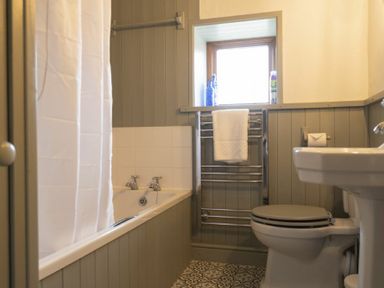 A bathroom with a bathtub and shower curtain a toilet and sink a towel rail under a window and tiled floor at Kittiwake Cottage Seahouses