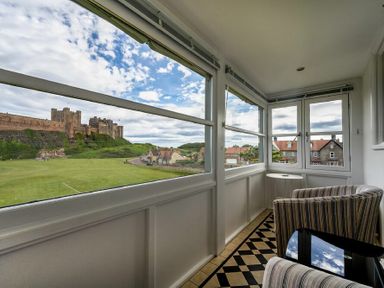An enclosed balcony with two striped chairs a small round table and large windows overlooking a castle and houses at Keepers View in Bamburgh