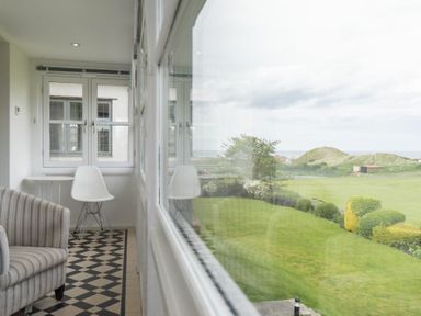 A narrow room with a striped armchair a white chair and windows overlooking a garden at Keeper's View in Bamburgh