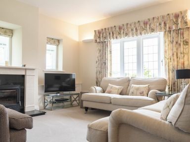 A living room with beige sofas floral curtains a television on a stand and a fireplace at Keepers View in Bamburgh