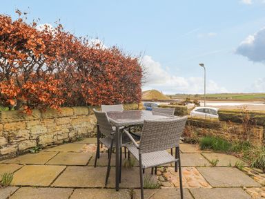 An outdoor patio with a glass table and four wicker chairs surrounded by a stone wall and hedge at The Beach House Alnmouth