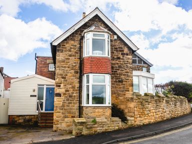 An exterior view of a stone house with bay windows and a blue door next to a stone wall on a street at The Beach House in Alnmouth