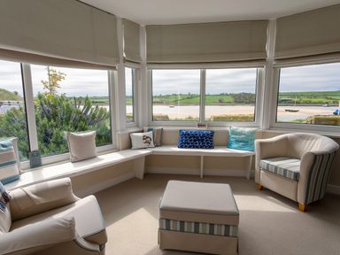 A seating area with armchairs a bench with cushions an ottoman and large windows overlooking water and greenery at The Beach House Alnmouth