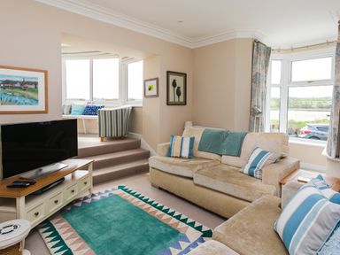 A living room with beige sofas and striped cushions a teal rug a television on a wooden stand and a small raised seating area at The Beach House Alnmouth
