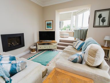 A living room with a beige sofa, a flat screen TV on a wooden stand, and a striped armchair in a sunlit seating area at The Beach House, Alnmouth