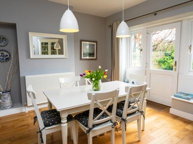 A dining room with a white table and six chairs with cushions flowers on the table and glass doors leading outside at The Beach House Alnmouth